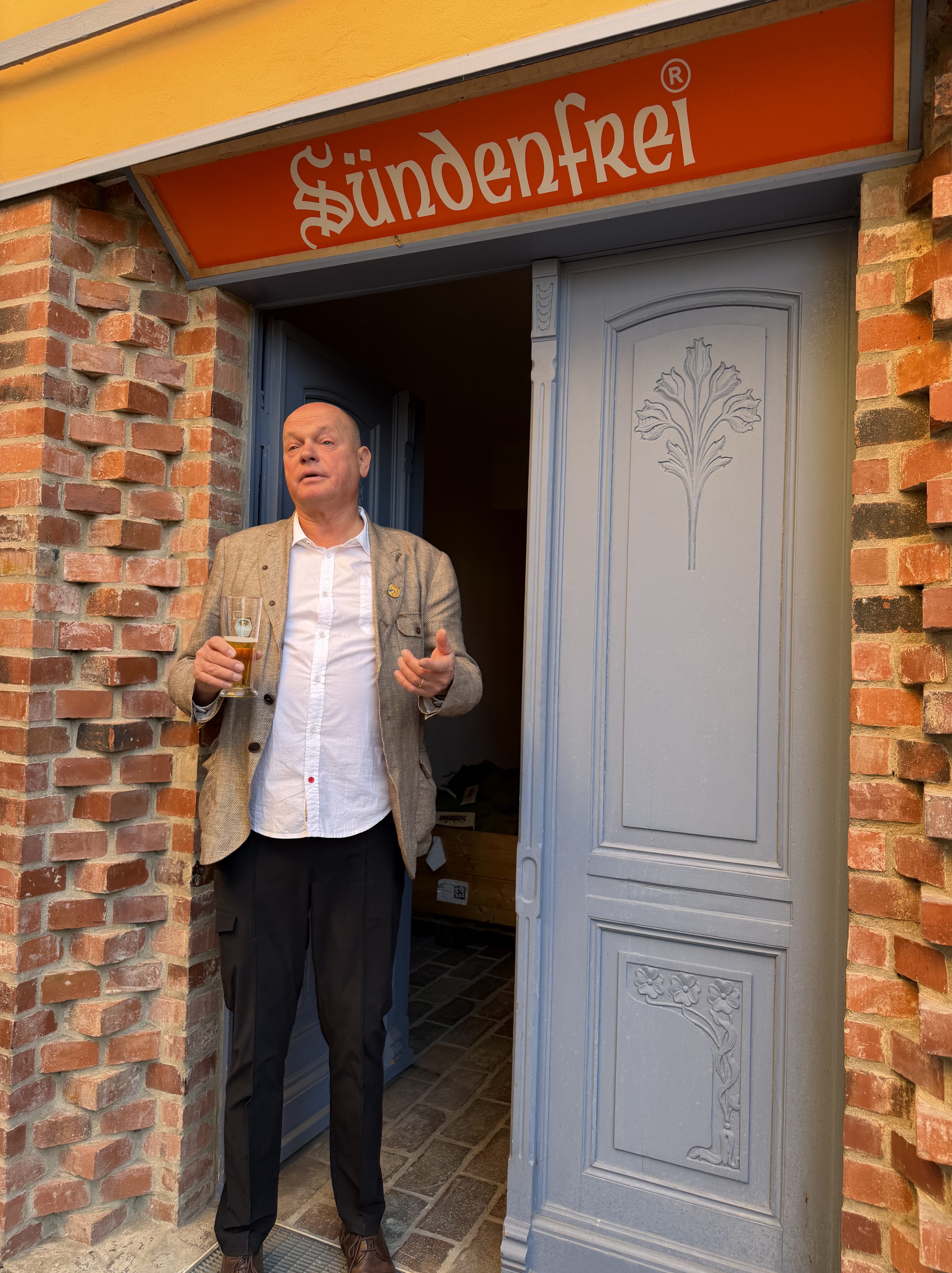 Ein Mann mit einem Getränk steht lächelnd am Eingang eines mit Ziegelsteinen verkleideten Gebäudes mit einem Schild "Sündenfrei".