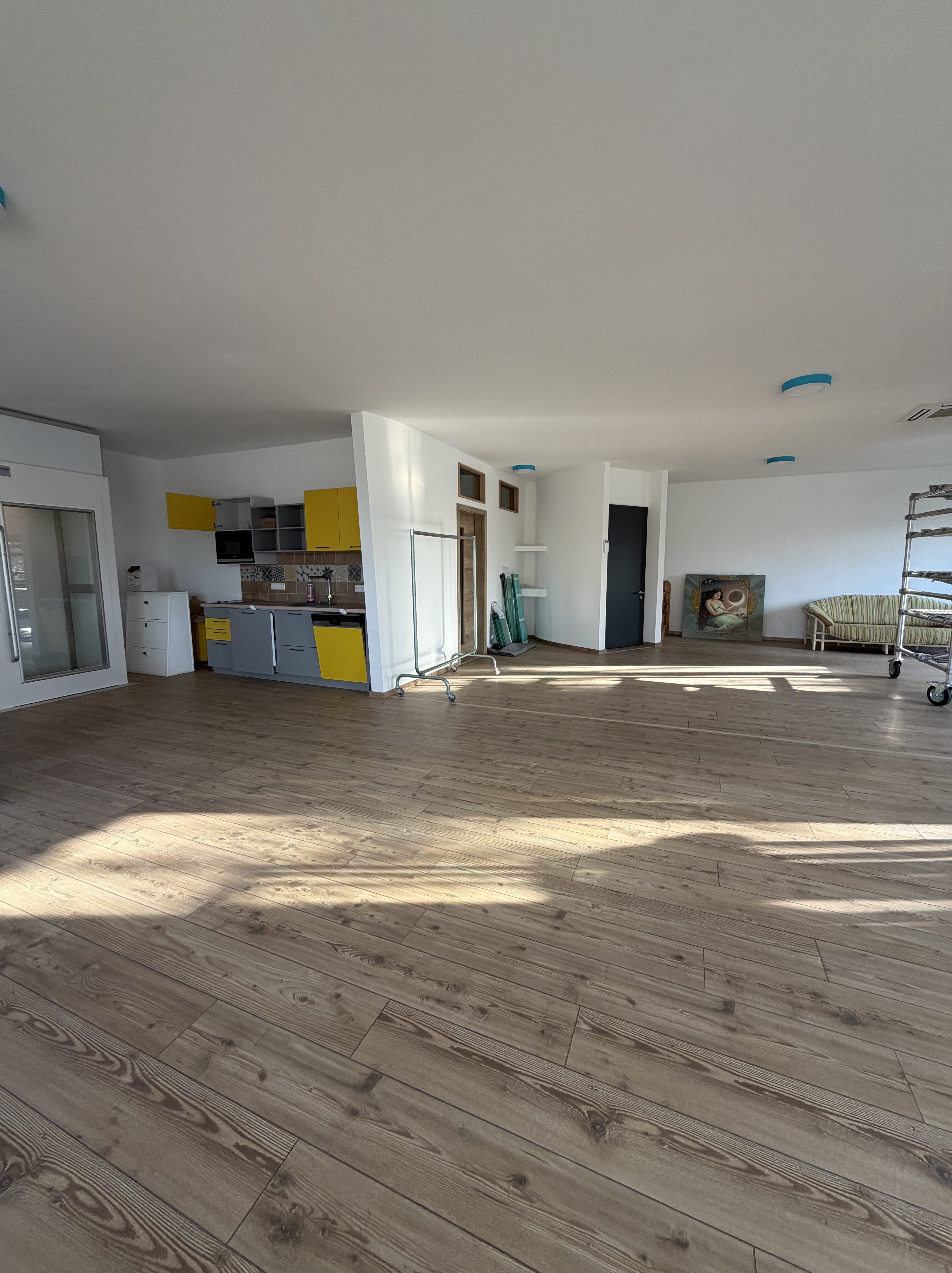 Heller, geräumiger Raum mit Holzfußboden und Küchenzeile in Gelb und Grau. Im Hintergrund stehen Möbel und ein Regal.