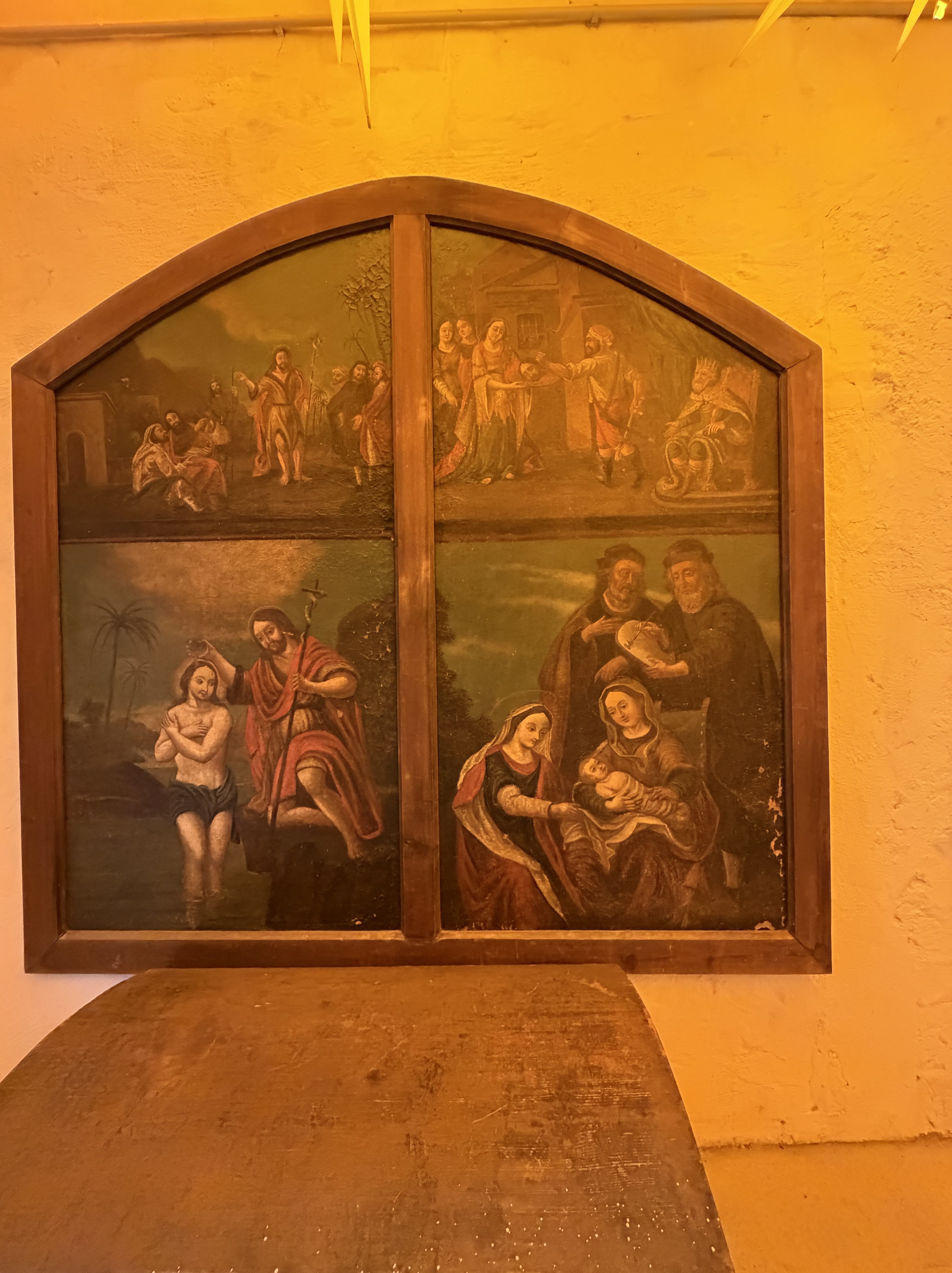 Ein historisches Gemälde in einem gebogenen Holzrahmen, unterteilt in vier Szenen mit religiösen Motiven, darunter Darstellungen von Heiligen und biblischen Ereignissen.