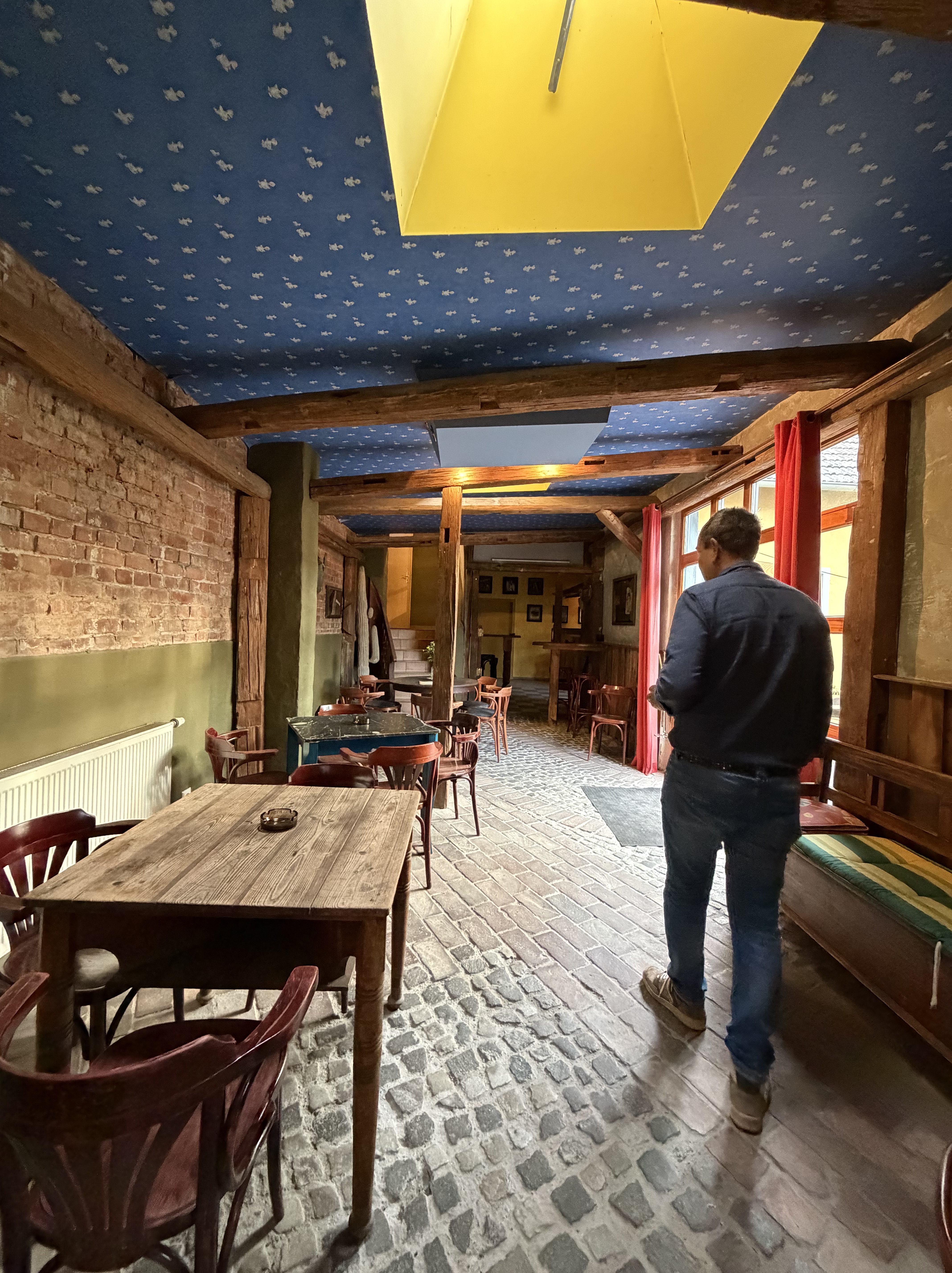 Ein gemütliches Restaurant mit Holztischen und -stühlen, einer Steinboden und farbenfrohem Wänden. Eine Person geht durch den Raum.