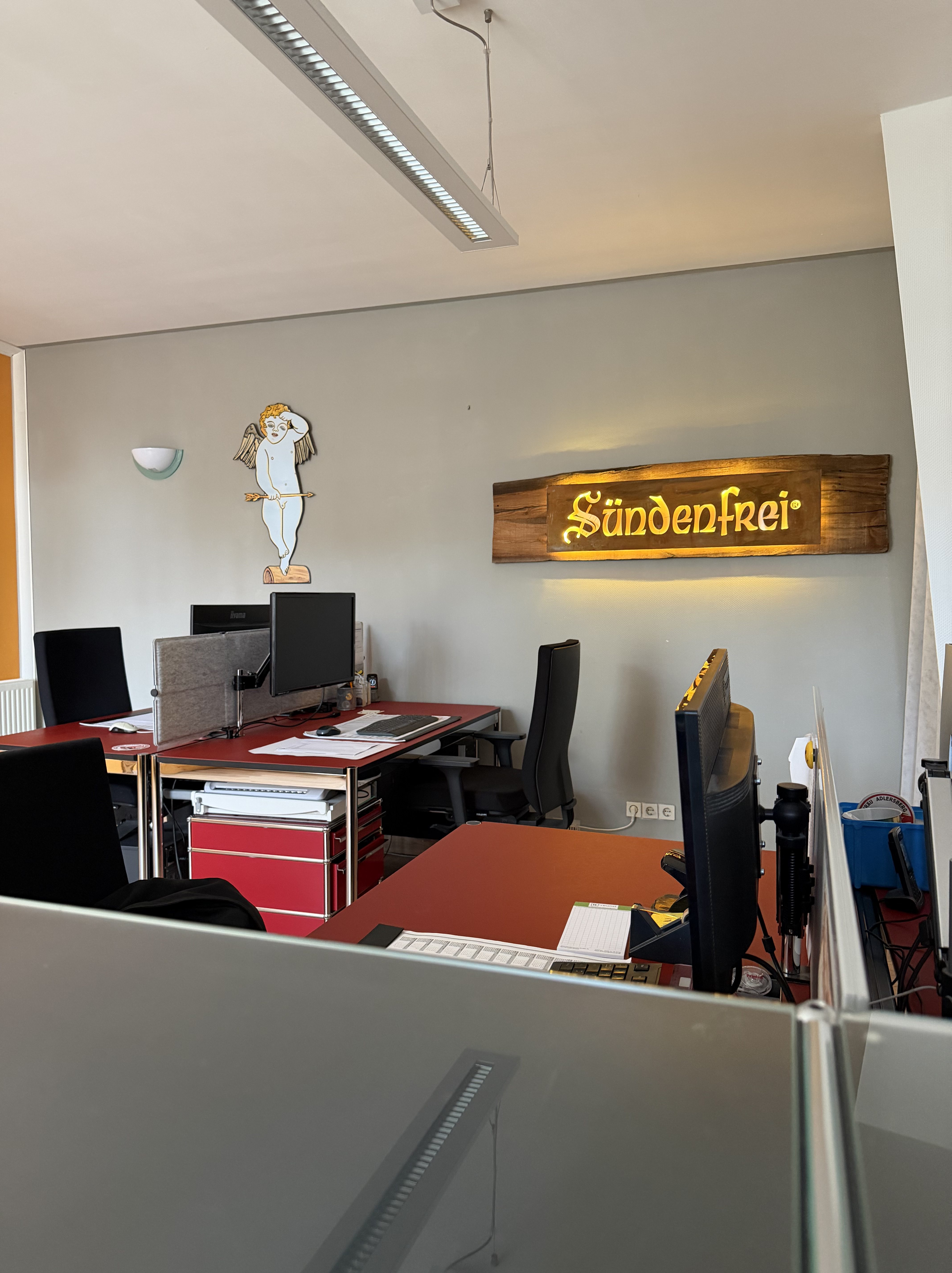 Ein modernes Büro mit roten Schreibtischen und mehreren Computern. An der Wand hängt eine bunte Figur und ein Holzschild mit der Aufschrift 'Sündenfrei'.