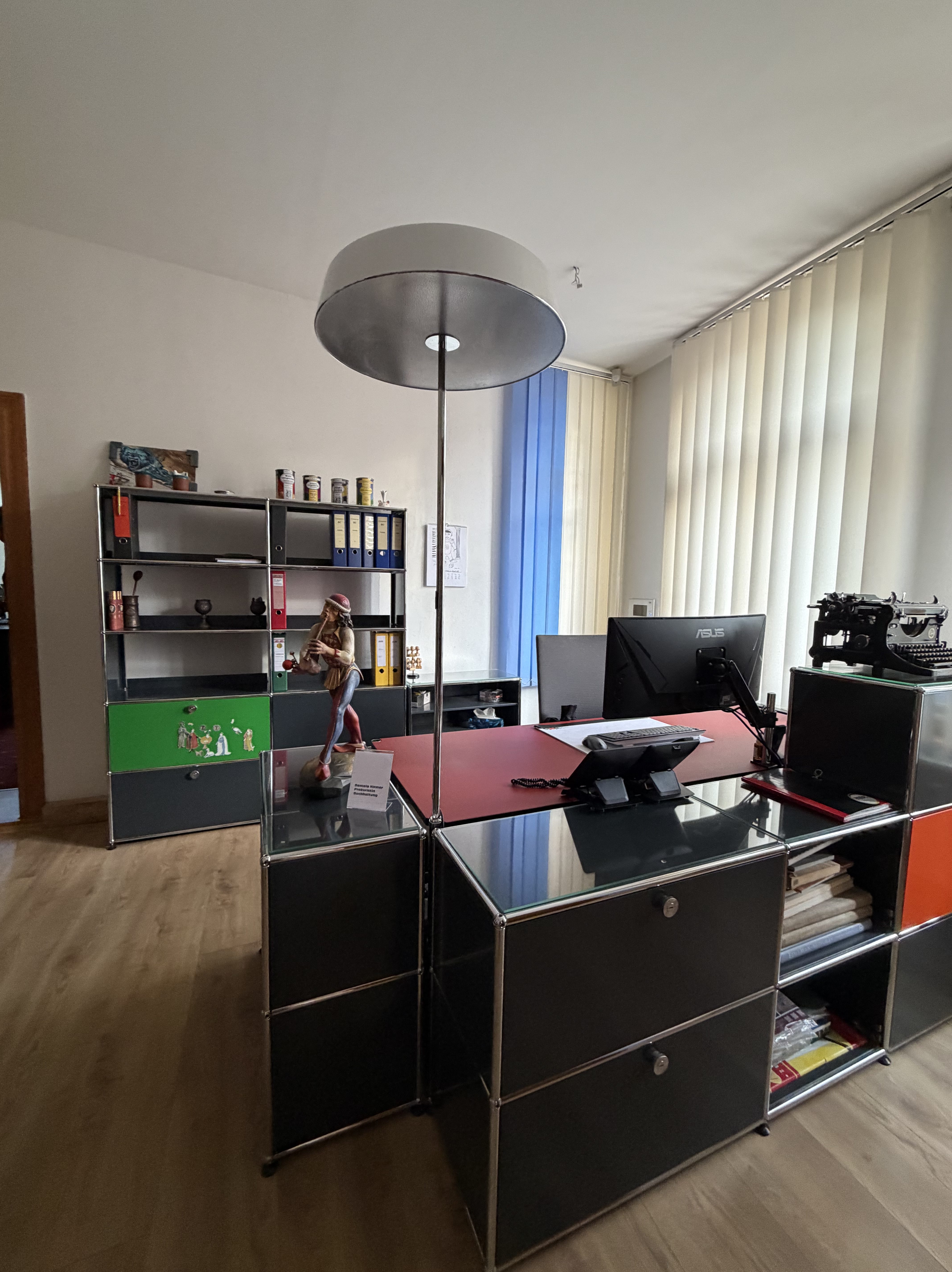 Ein modernes Büro mit einem Schreibtisch, einem Computer, einem Telefon und verschiedenen Regalen. Auf dem Schreibtisch steht eine Lampe und eine Figur. Die Wände sind mit Jalousien und Dekorationen geschmückt.