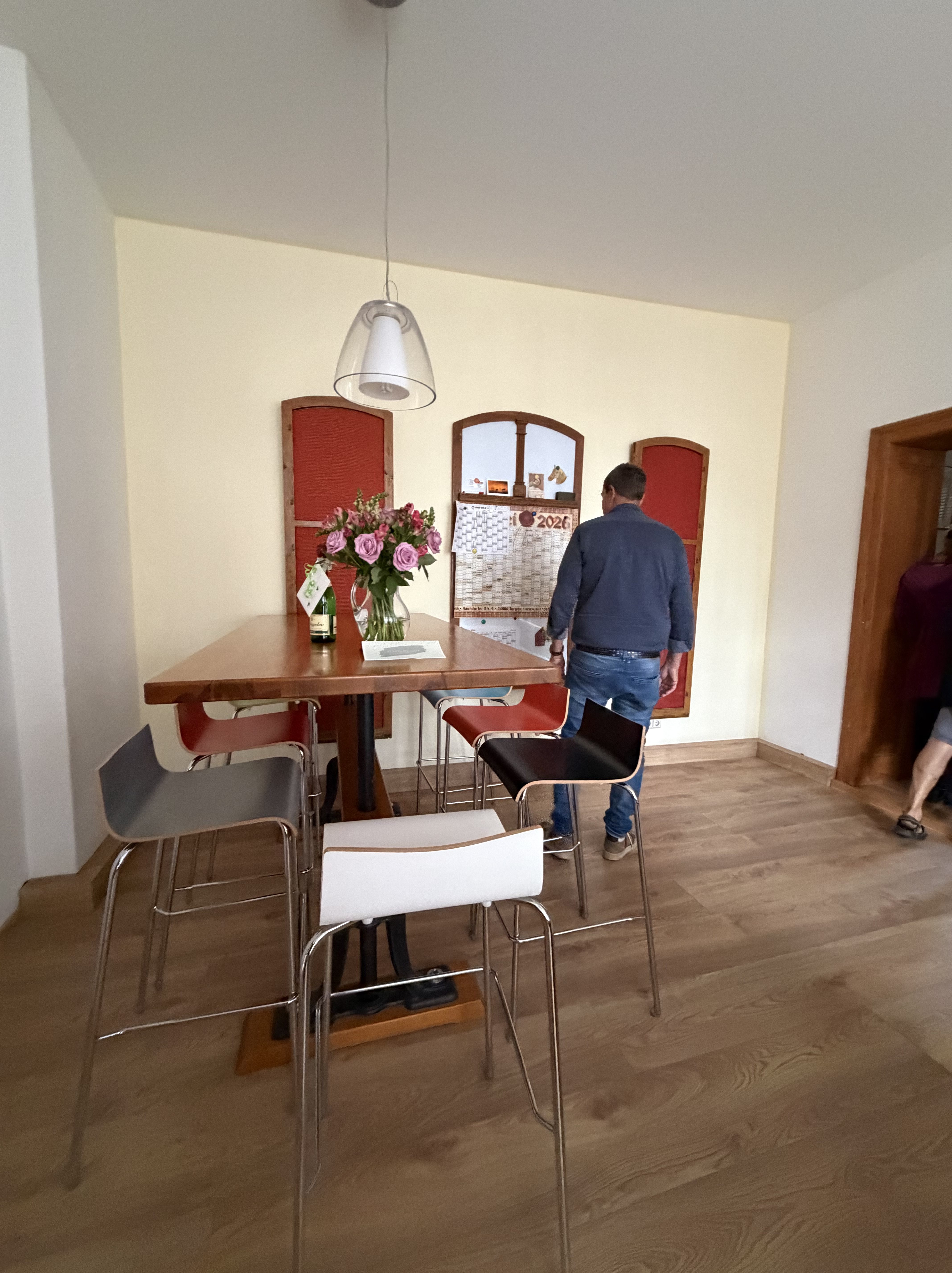 Ein moderner Essbereich mit einem Holzstisch und bunten Stühlen. Auf dem Tisch steht eine Vase mit Rosen und daneben liegt ein Kalender. Eine Person, die einen blauen Pullover trägt, steht am Tisch und schaut in Richtung einer Wand mit Fensterläden.