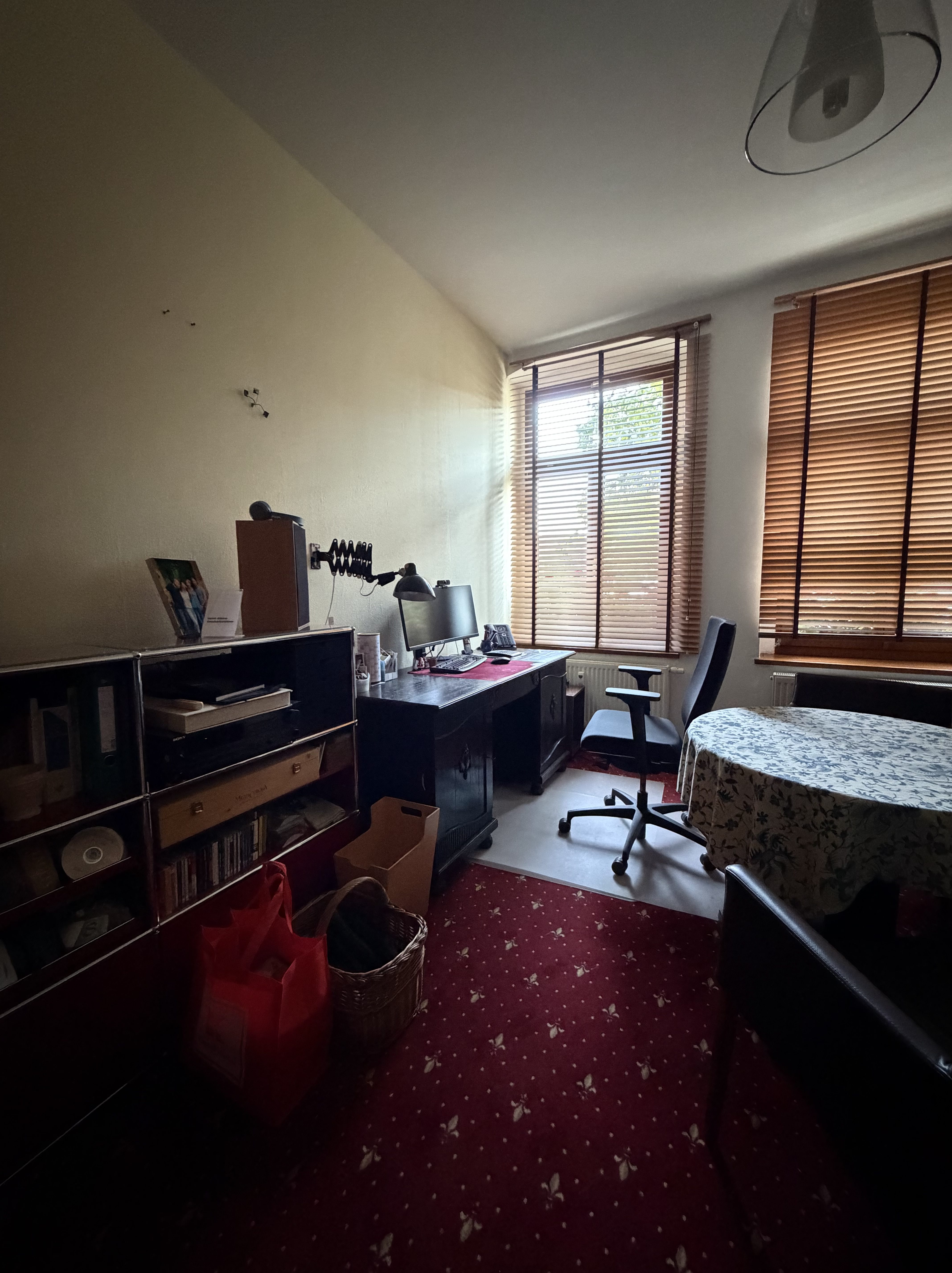Ein Arbeitszimmer mit einem Schreibtisch, einem Computer, einem Bürostuhl und einem Tisch mit floralem Tischdecken. Im Raum stehen Regale mit Büchern und Aufbewahrungskörben. Das Licht fällt durch Jalousien an einem Fenster.