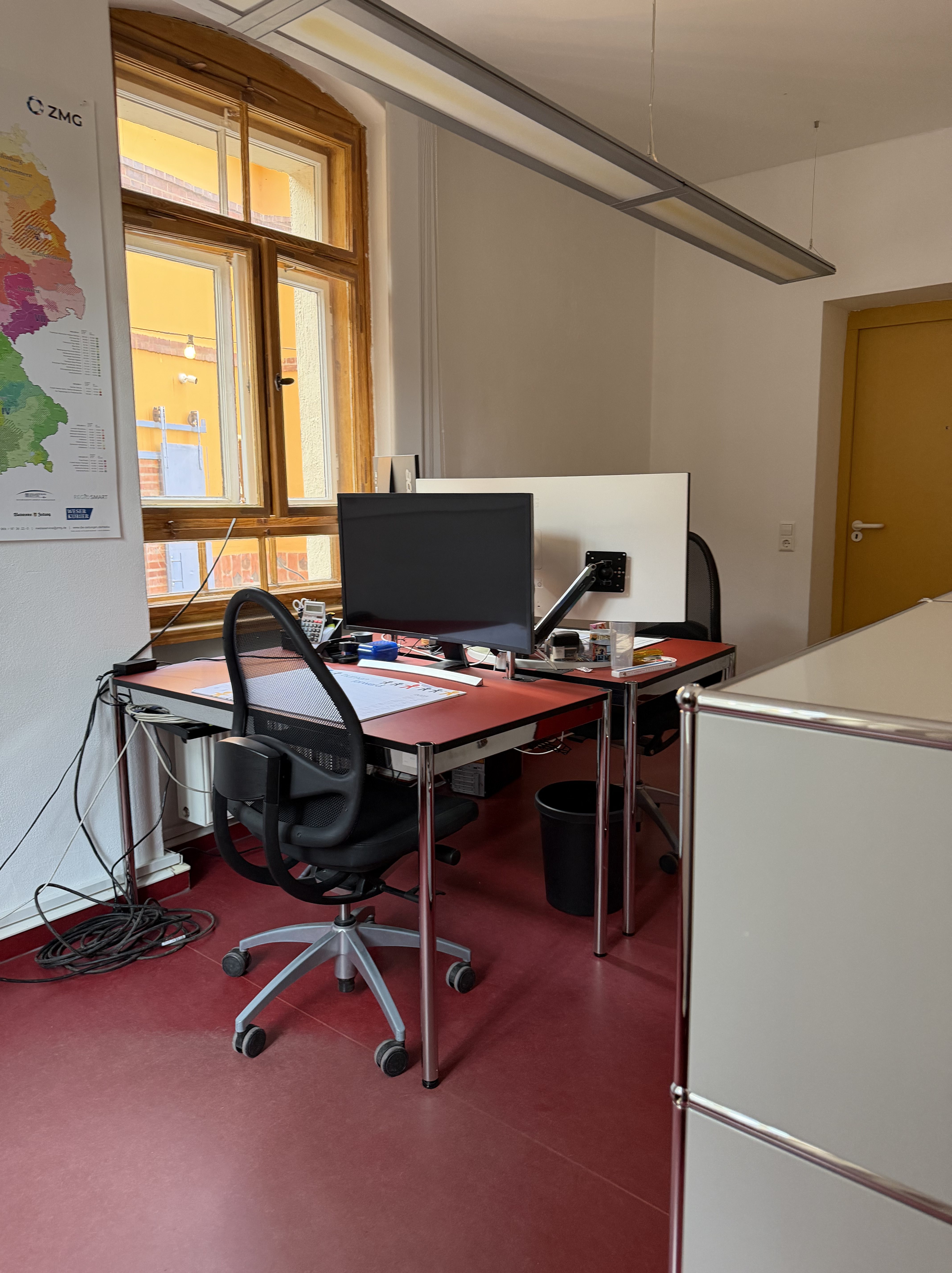 Ein modernes Büro mit zwei Schreibtischen, einem Computerbildschirm und einem ergonomischen Bürostuhl. Im Hintergrund ist ein Fenster mit Blick auf eine farbige Wand. Auf dem Tisch liegen verschiedene Büroutensilien und ein müllbehälter.