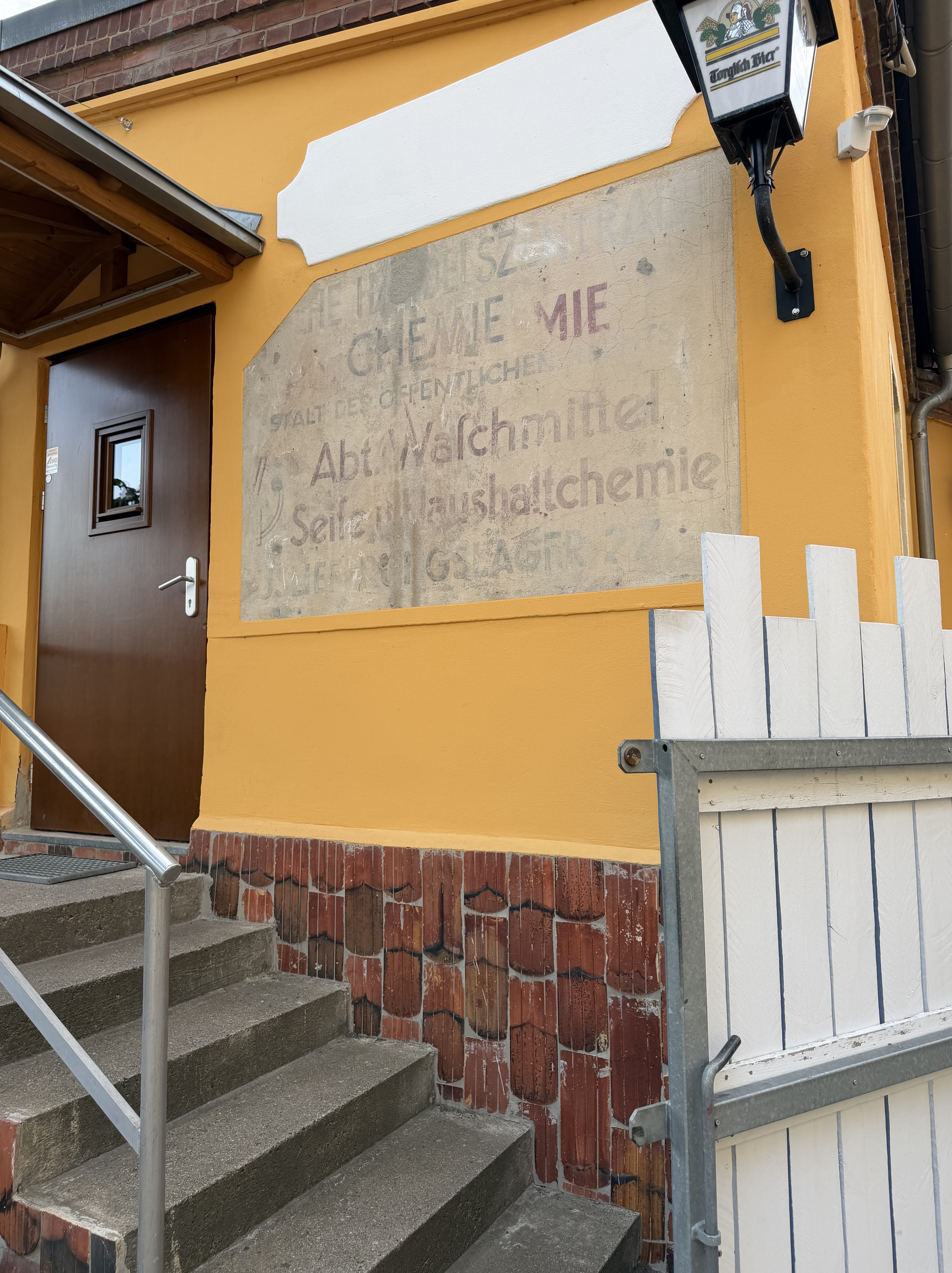 Eingang zu einem Gebäude mit einer alten, teilweise verwitterten Werbetafel für chemische Produkte und einer braunen Holztür, umgeben von gelber Wandfarbe und einer weißen Holzlattenzaun.