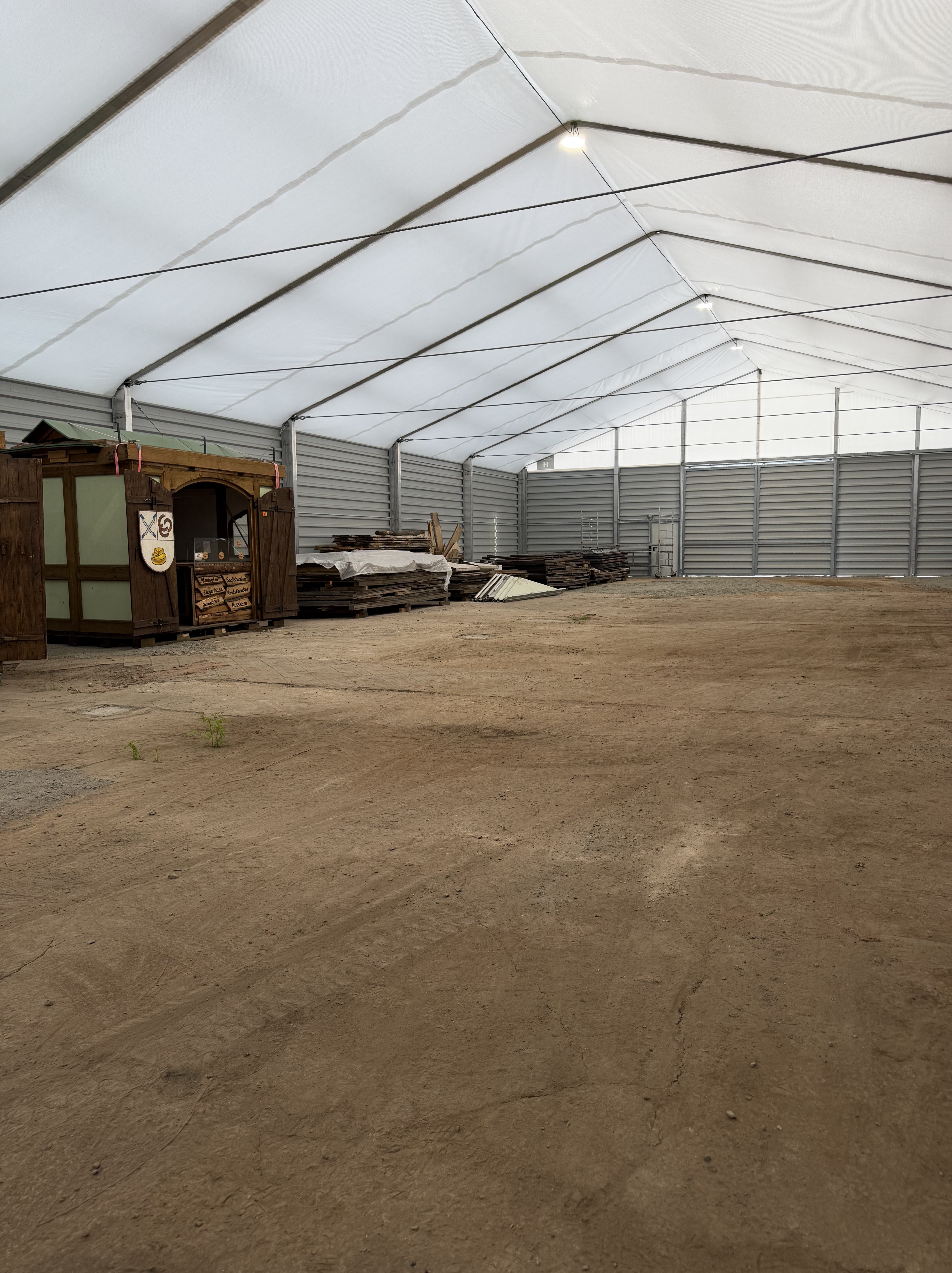 Eine große, leere Lagerhalle mit einem Zeltoberteil, in dem einige Holzstapel und eine kleine hölzerne Struktur im Hintergrund zu sehen sind. Der Boden ist staubig und unbefestigt.