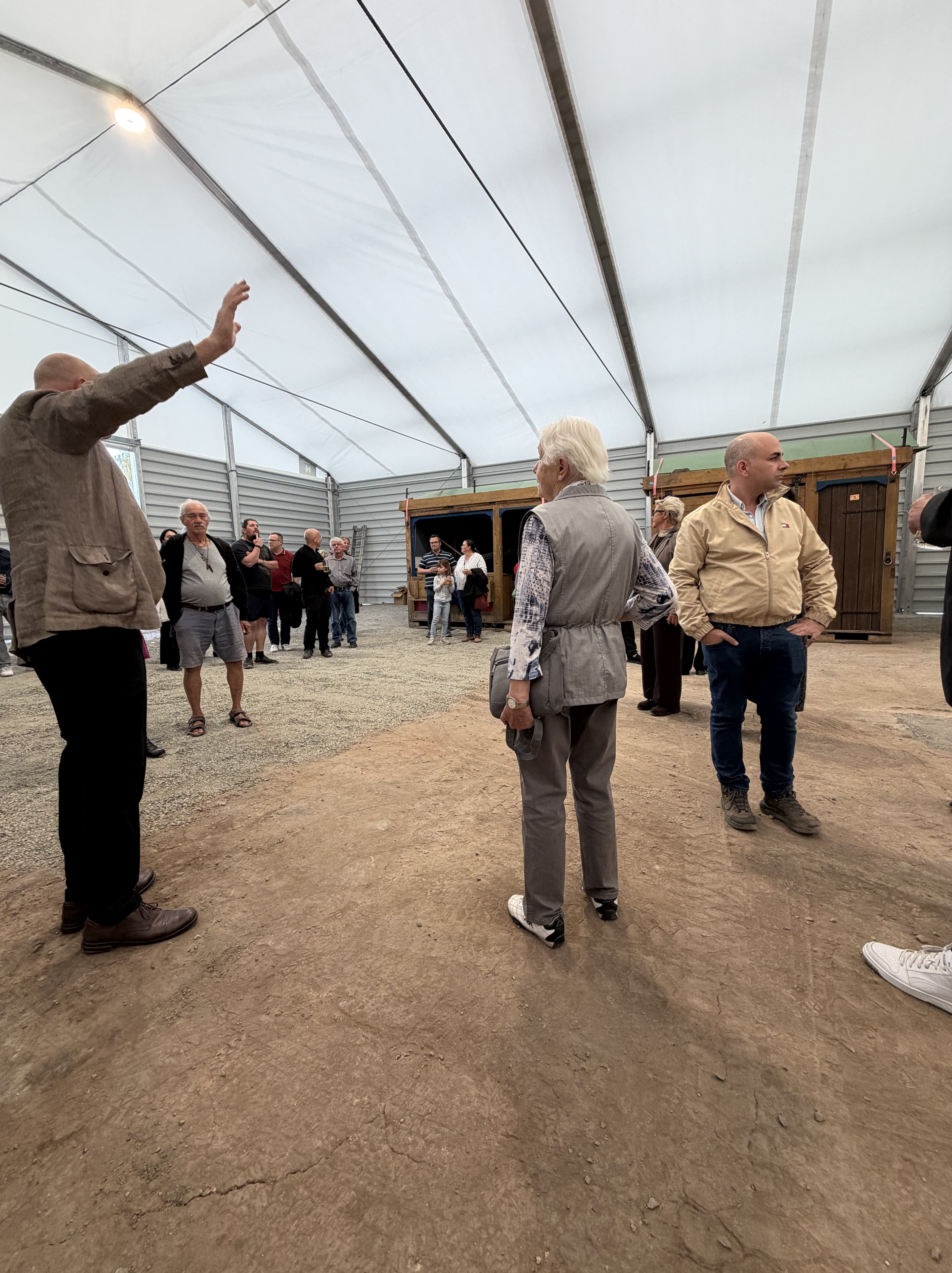 Eine Gruppe von Menschen steht in einem großen Zelt mit einem hellen Dach. Eine Person steht mit erhobener Hand und spricht zu den anderen, die interessiert zuhören oder sich unterhalten.