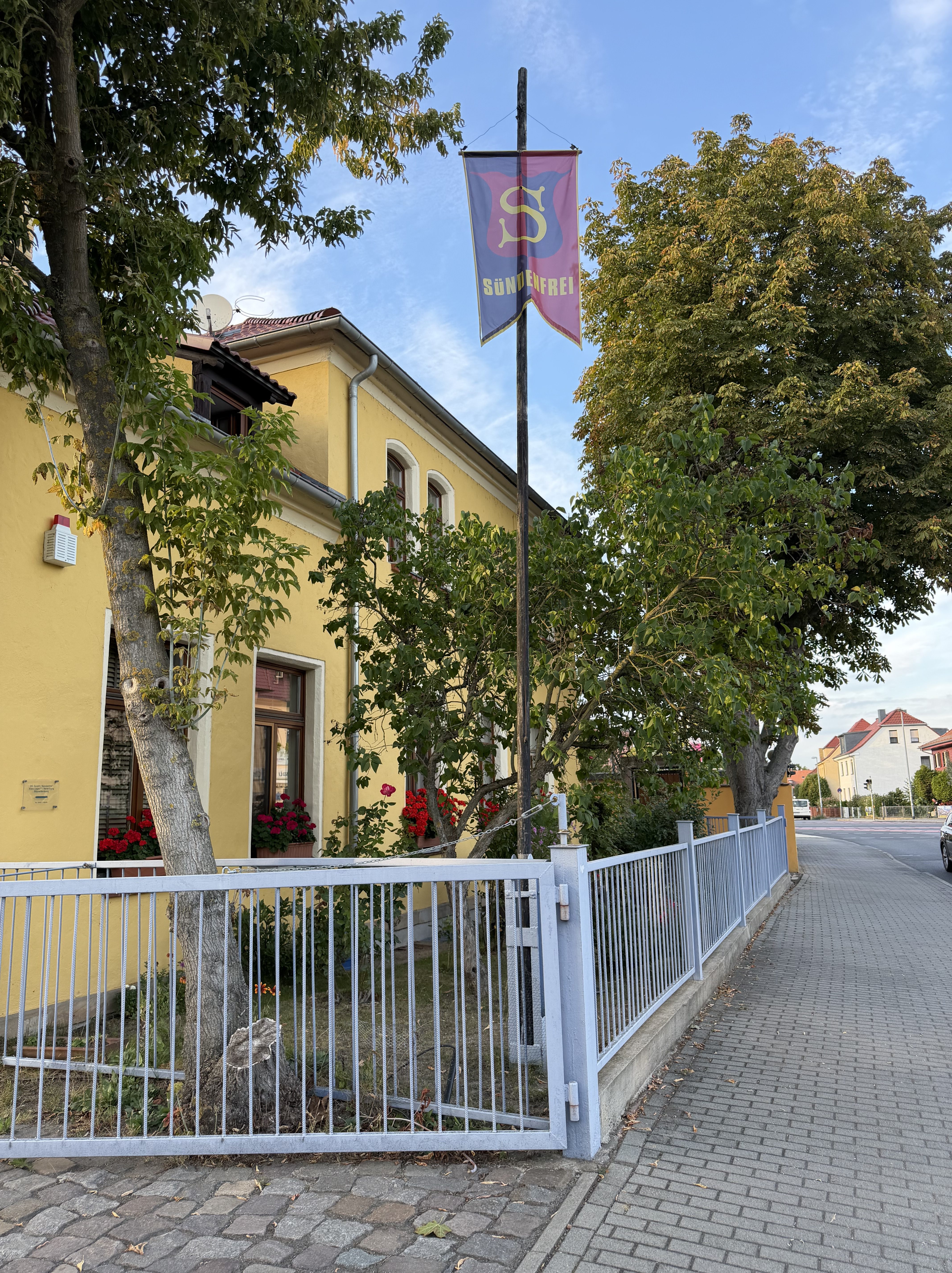 Gelbes Gebäude mit einem Schild, das 'Sündenfrei' anzeigt, umgeben von Bäumen und Blumen.