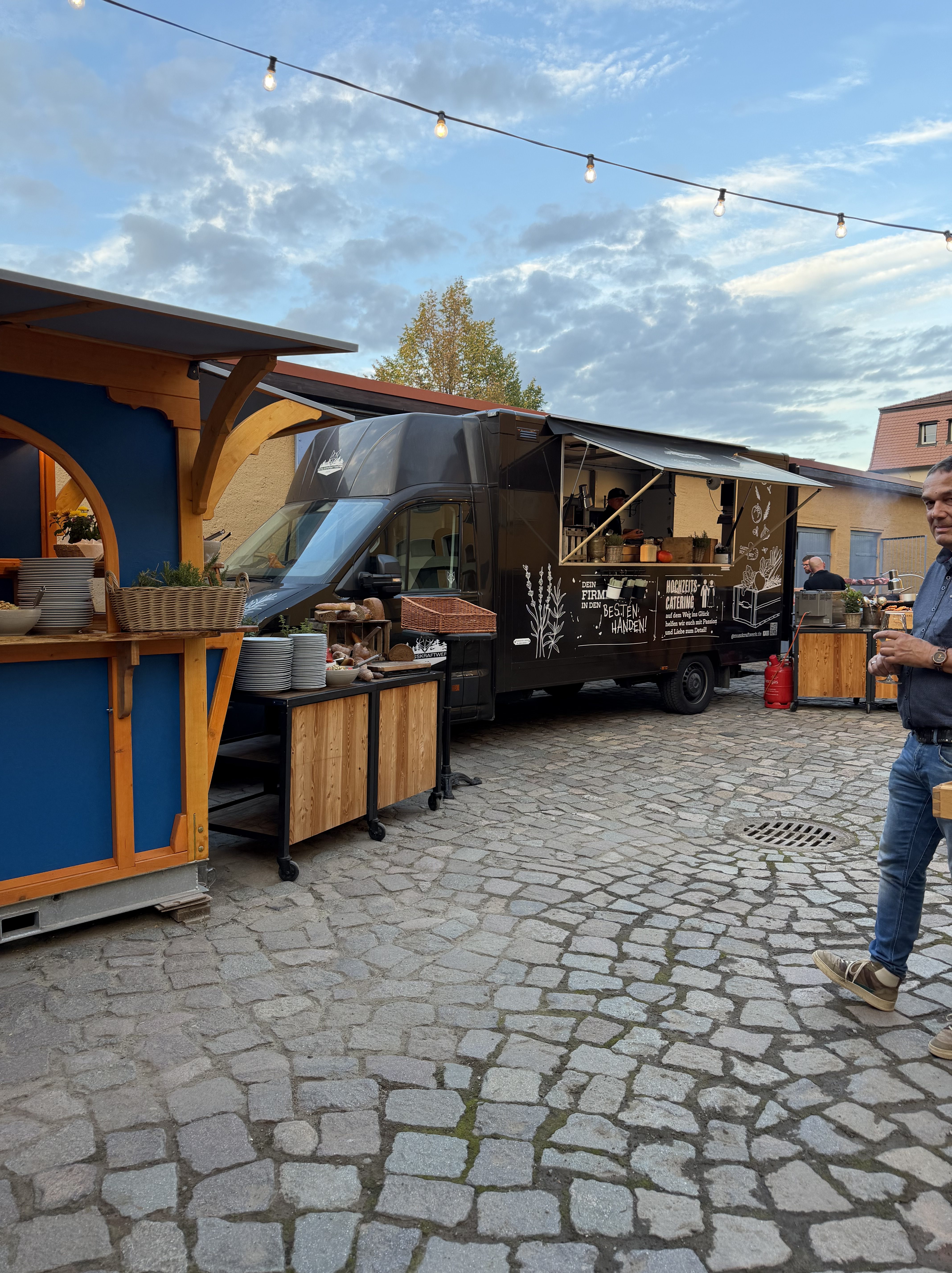 Ein mobiler Foodtruck auf einem gepflasterten Platz, umgeben von Verkaufsständen. Der Himmel ist bewölkt und es hängen Lichterketten über dem Bereich.