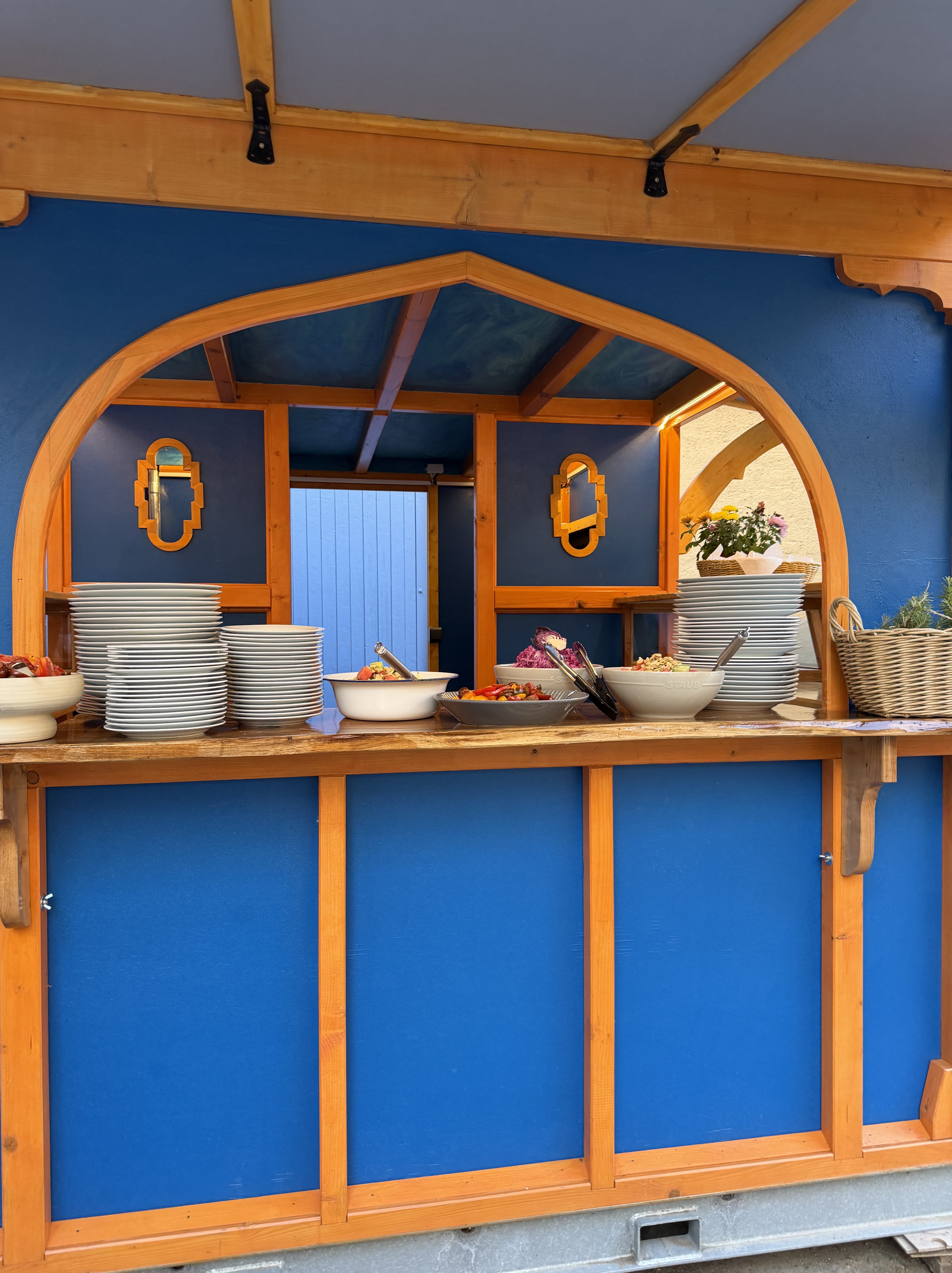 Ein Buffetstand mit blauen Wänden, Holzrahmen und mehreren gestapelten Tellern. Verschiedene Salate und Früchte in Schüsseln sind auf der Theke angerichtet, mit dekorativen Spiegeln an den Wänden.