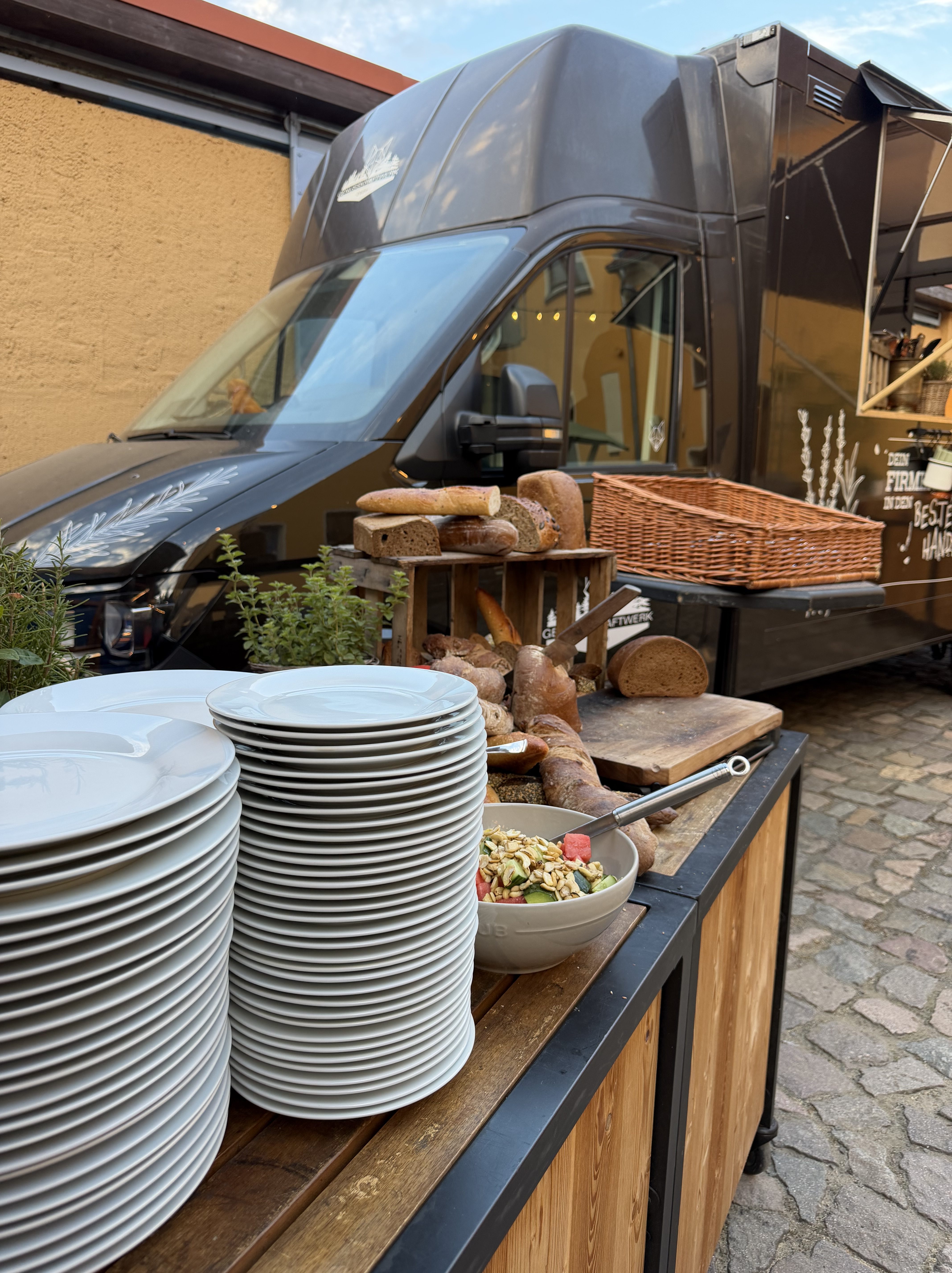 Ein Food Truck mit einem Angebot an frischem Brot, gestapelten Tellern und einer Schüssel mit Müsli und Obst auf einem rustikalen Holzstand.