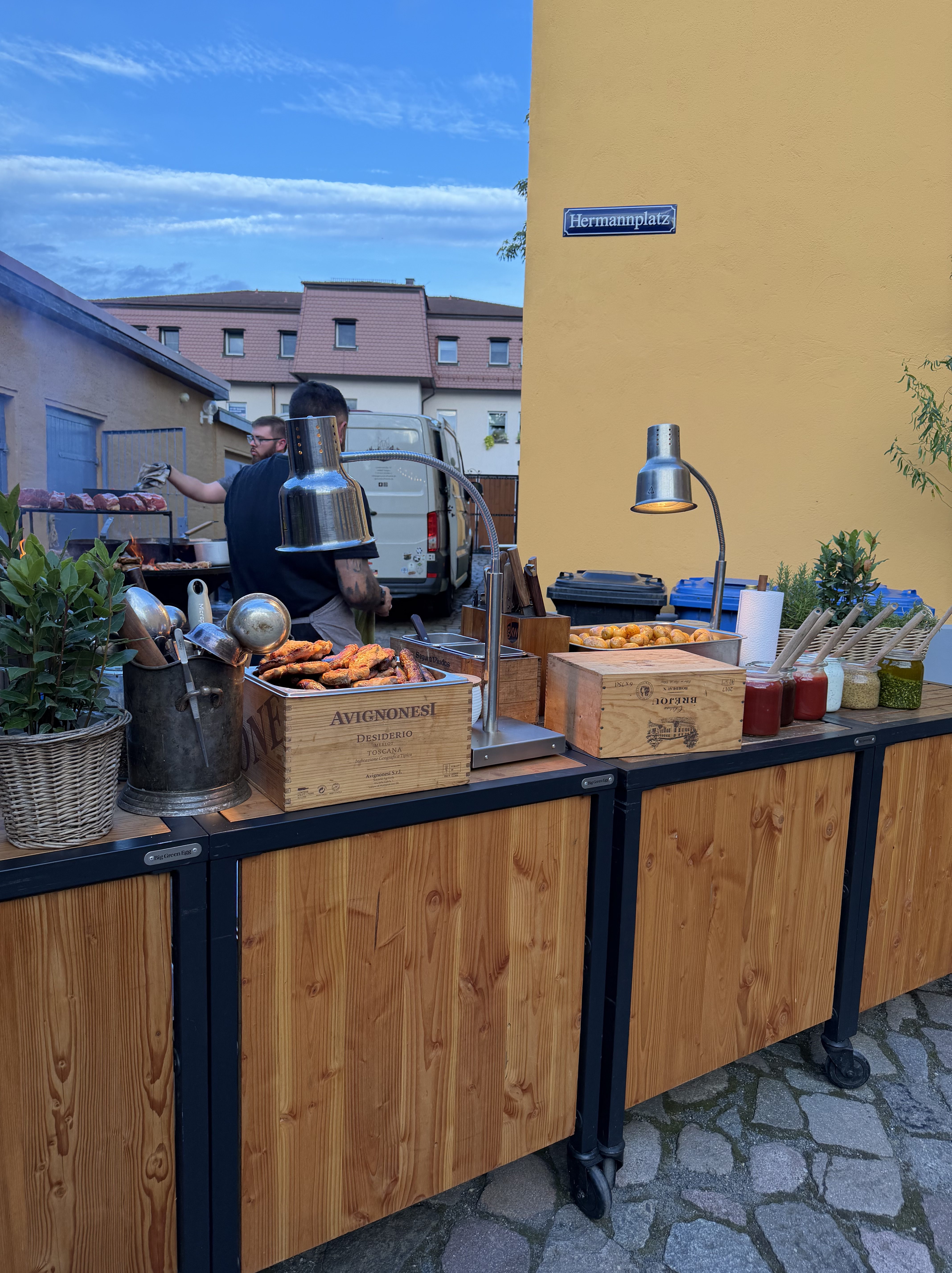 Eine Grillstation im Freien mit verschiedenen Speisen auf Holztischen, darunter mariniertes Fleisch und Beilagen. Im Hintergrund ist ein Mann zu sehen, der am Grill arbeitet. Ein Straßenschild zeigt den Namen "Hermannplatz".