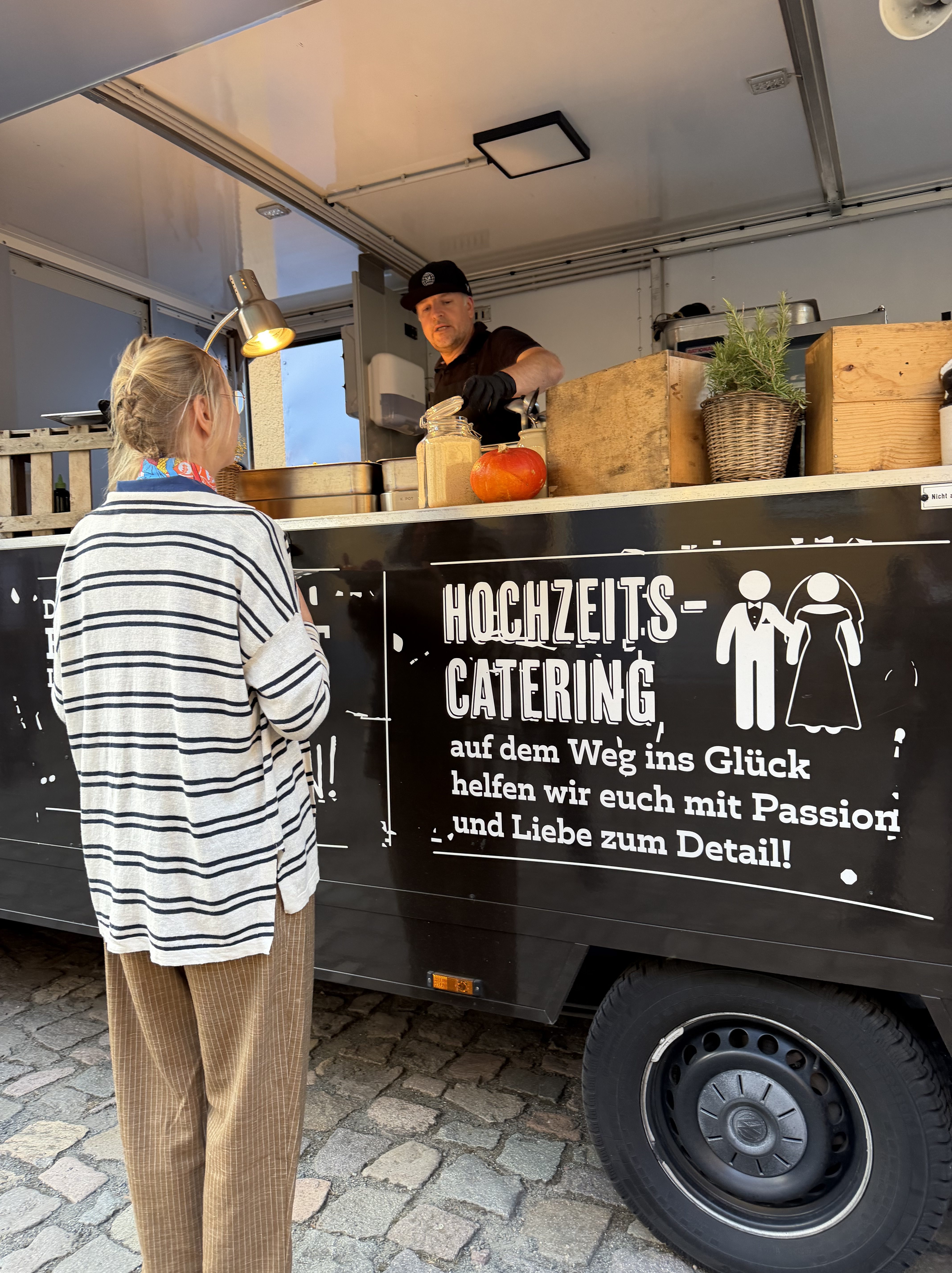 Eine Person steht vor einem Catering-Foodtruck, der Hochzeitcatering anbietet. Der Verkäufer, der einen schwarzen Hut trägt, bedient die Kundin, die ein gestreiftes Oberteil und eine farbige Halskette trägt. Auf dem Truck sind Details zur Hochzeit und Catering zu sehen.