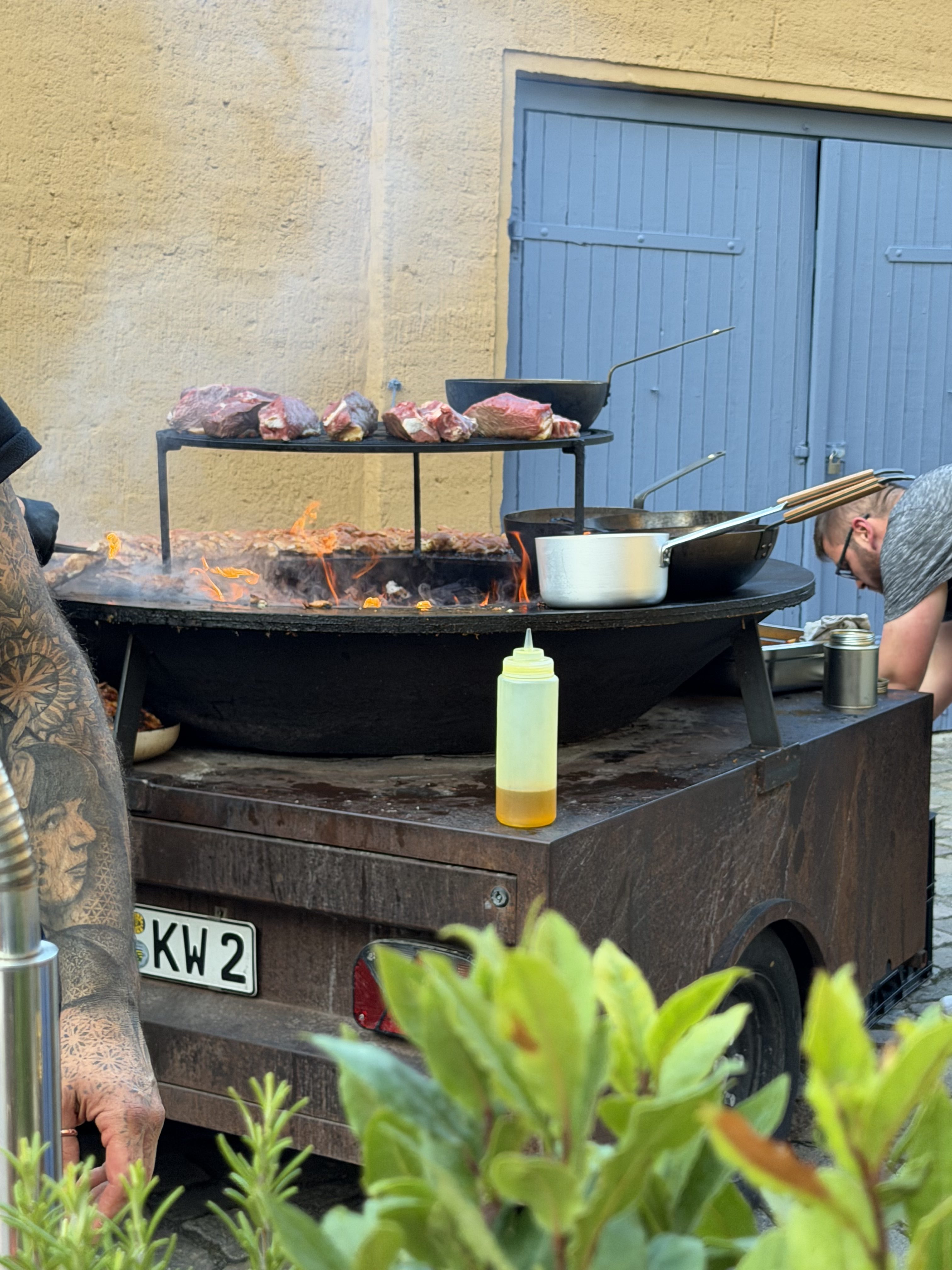 Ein Grillstand im Freien mit rauchendem Kohleofen, auf dem Fleischstücke braten. Im Hintergrund sind Küchenutensilien und ein Koch zu sehen.