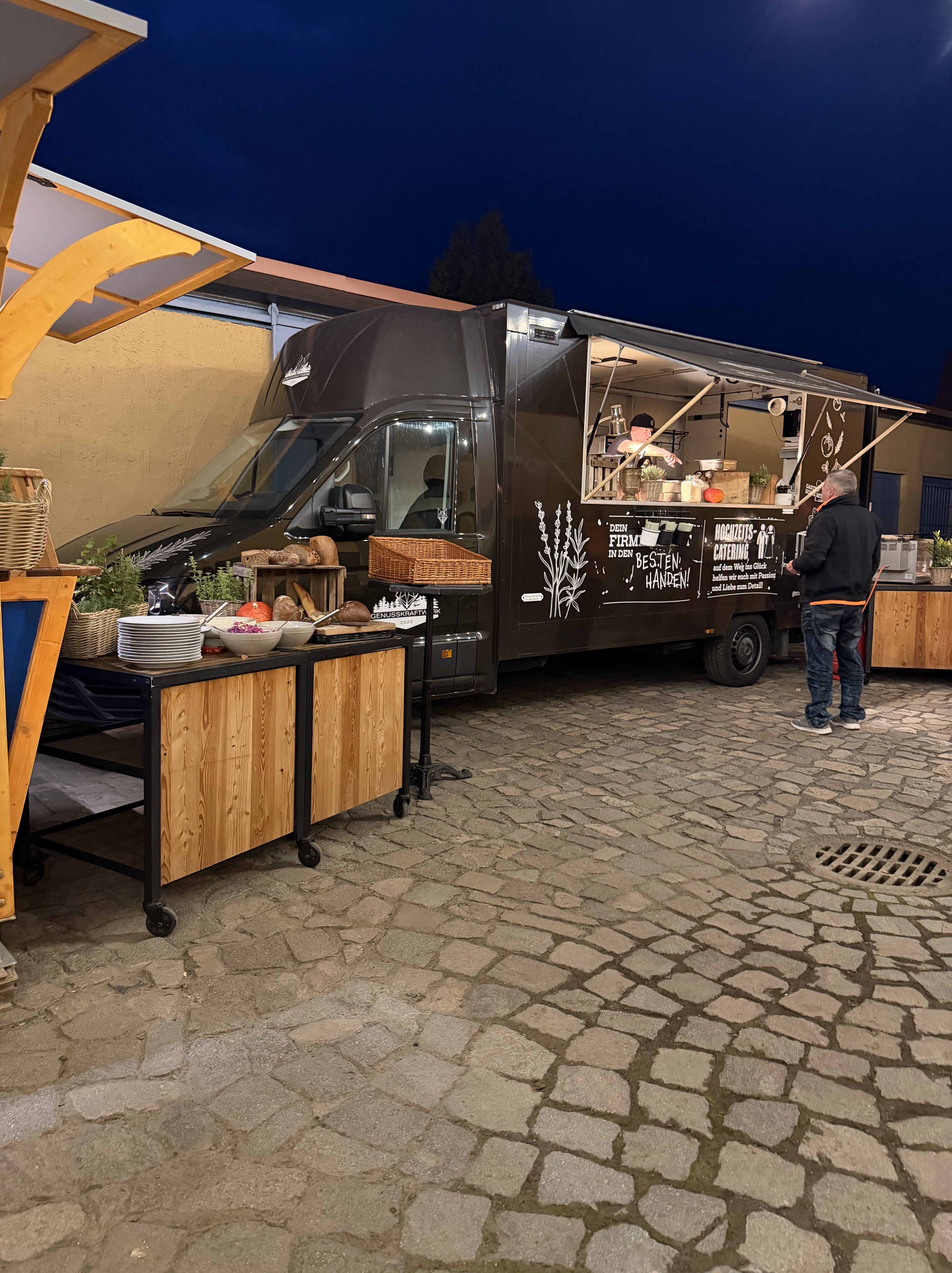 Ein Foodtruck mit einem offenen Fenster, von dem aus Essen verkauft wird. Vor dem Truck stehen Holzstände mit verschiedenen Lebensmitteln und Geschirr. Die Umgebung ist abends, im Hintergrund ist der dunkle Himmel zu sehen.