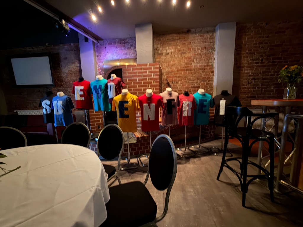 Bunte T-Shirts auf Ständern mit den Buchstaben 'S, E, I, N, F, R, E', im Hintergrund eine Backsteinwand.